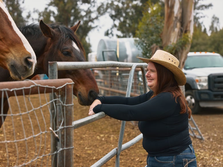A cowgirl wearing the Outback Trading Co Palm Collection Santa Fe Straw Hat looking at two horses in a field