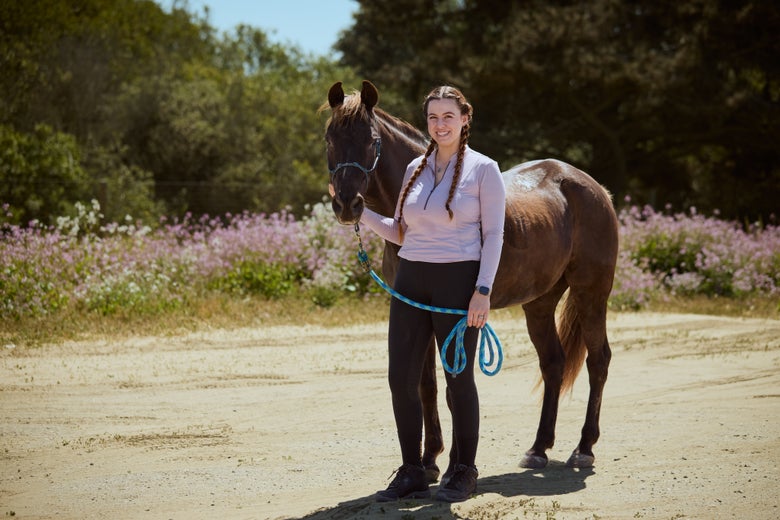 A rider wearing the Ariat Eos 2.0 Knee Patch Tights standing with her horse in front of a field of purple flowers.