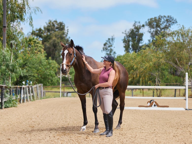 A rider wearing the Mountain Horse Diana Full Seat Breeches petting her bay horse in an arena.