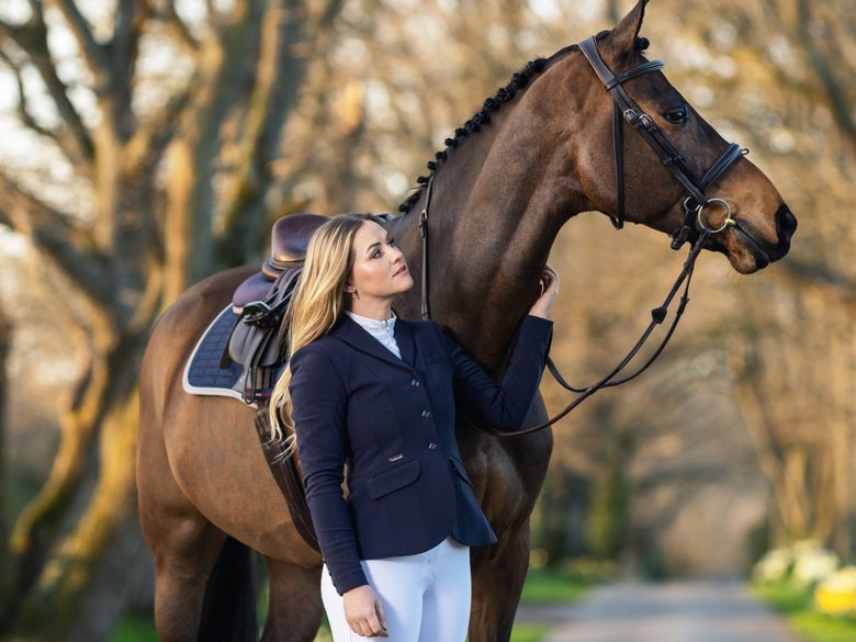 Woman standing next to her bay eventing horse, wearing the LeMieux Dynamique Show Jacket. 