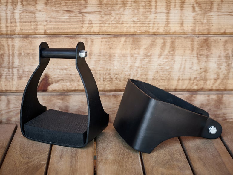 The Tough 1 Angled Aluminum Endurance Trail Stirrups sitting on a wooden floor against a wooden barn. 