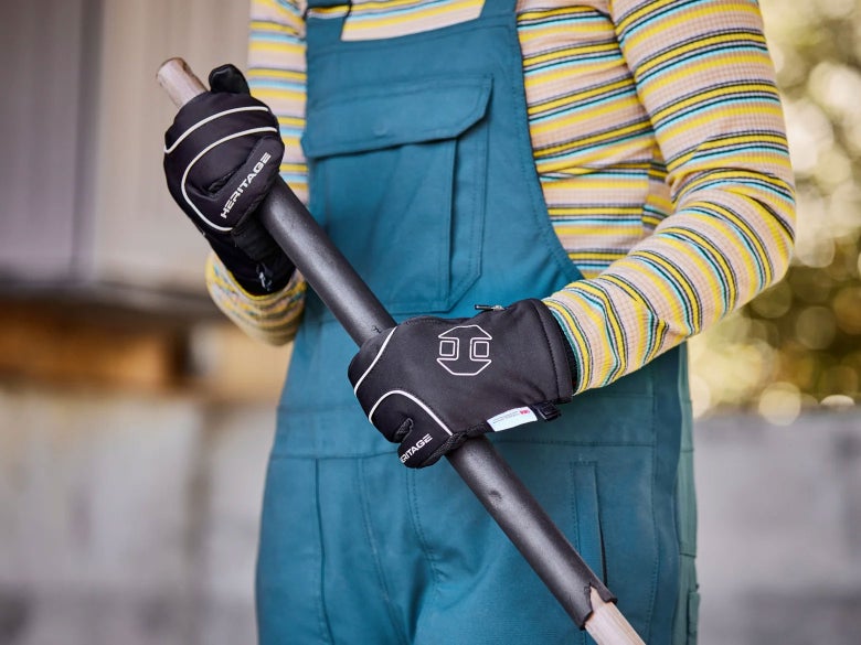 Woman in a striped long sleeve shirt and overalls, holding a muck fork with the Heritage Arctic Winter Riding Mitten Gloves on.
