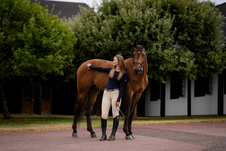 Girl wearing a TKEQ Equestrian Sun Shirt in black, standing next to her bridled bay horse.