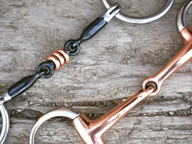 Close up image of a copper bit and sweet iron bit laying side-by-side on a piece of wood.