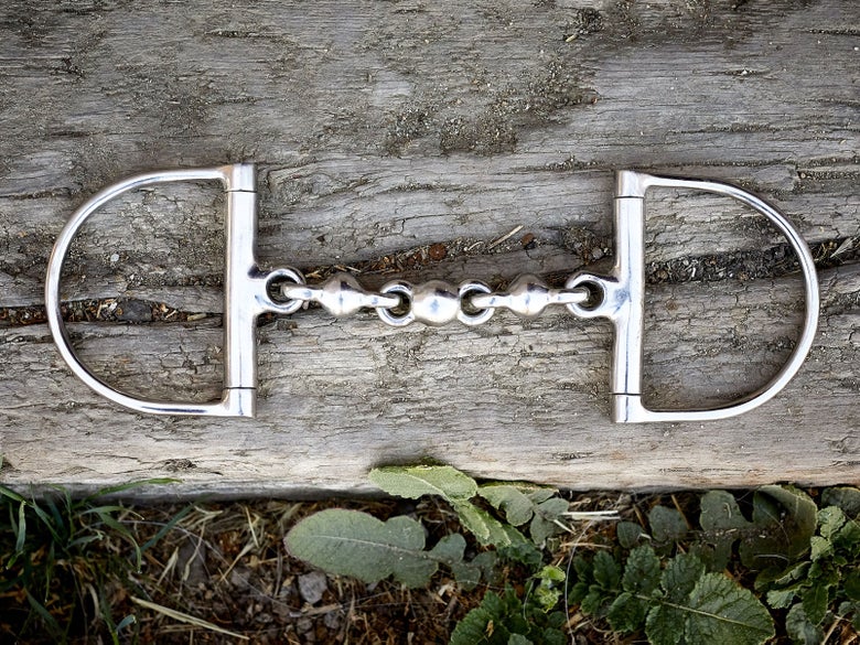 A horse bit with D-ring cheekpieces, laying flat on a piece of wood. 