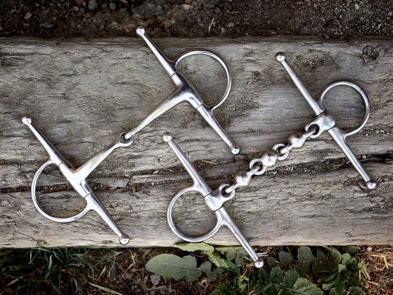 Two horse bits with full-cheek cheekpieces, laying side-by-side on a piece of wood. 