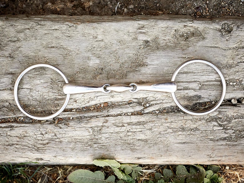 A horse bit with loose ring cheekpieces, laying flat on a piece of wood. 