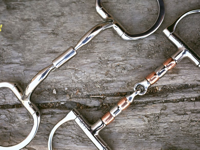 Close up image of two horse bits side-by-side with a roller mouthpieces, laying flat on a piece of wood. 