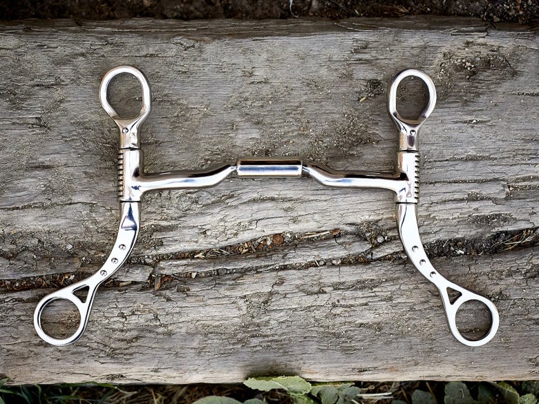 A horse bit with shank cheekpieces, laying flat on a piece of wood. 