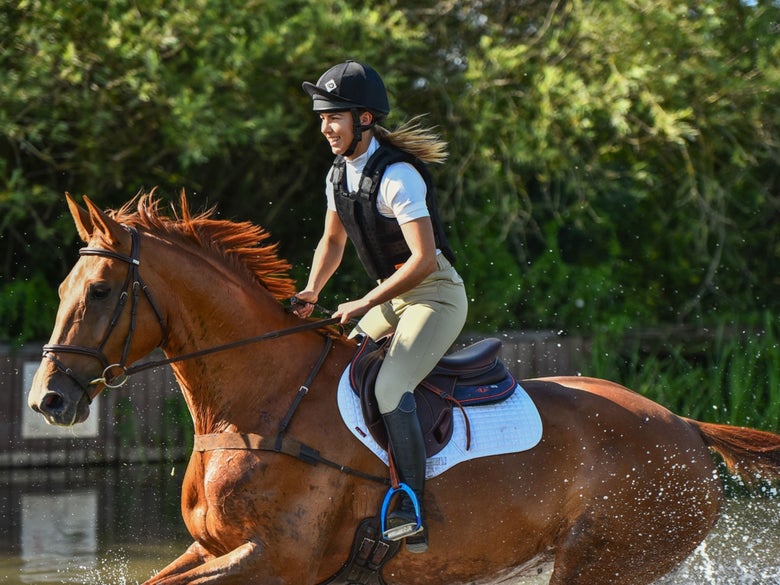 Lady wearing a Charles Owen helmet, riding her chestnut horse through the water on a cross country course