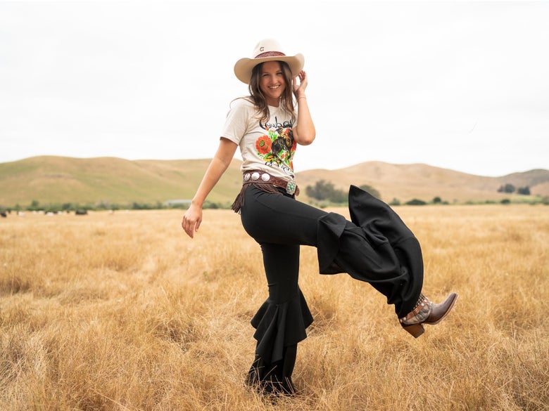 Lady wearing a fun western inspired outfit, kicking up her cowboy boots in a field