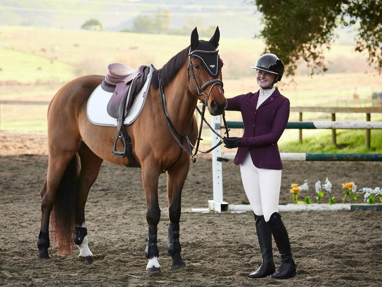 A rider in white breeches and a maroon show coat appropriately dressed for the show jumping ring, holding the reins of her tacked up bay horse. 