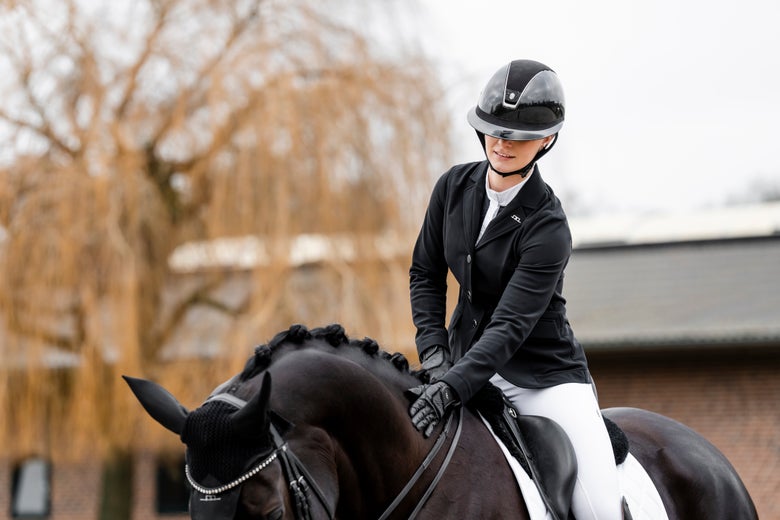 A rider petting her dark bay horse while wearing the AA Horseware Platinum Motionflex Pro Jacket.