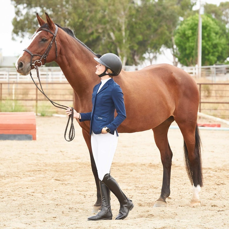 A rider dressed for the show jumping ring in a blue show coat and white breeches standing with a bay horse.