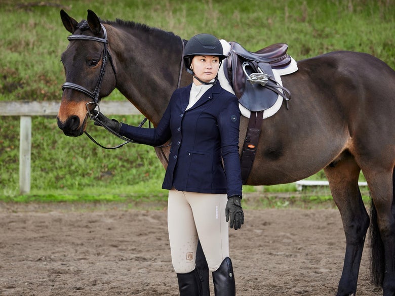 Rider wearing tan breeches and a navy show coat in appropriate hunter ring attire standing with her tacked up dark bay horse.