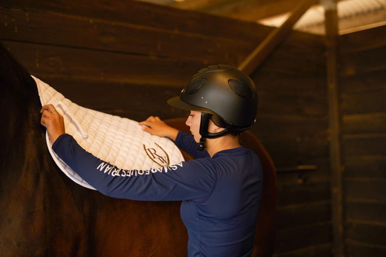 Woman placing a white english saddle pad on a bay horse in cross ties.