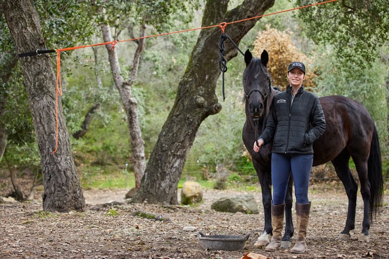 Gillian standing next to her black mare, tied on the Tough 1 high line with a feed pan on the ground. 