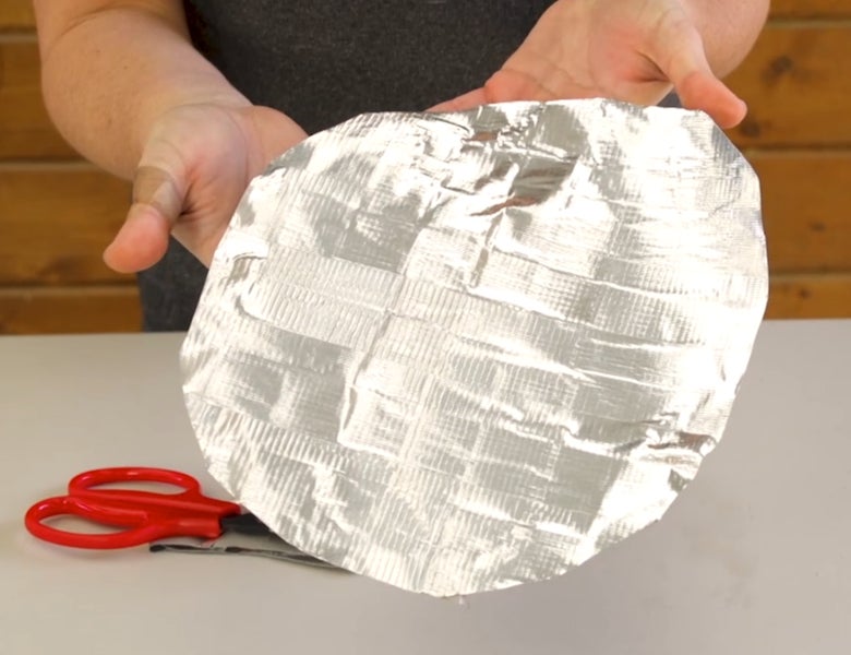 A woma's hands holding up a duct tape hoof boot with red scissors on a plastic table. 