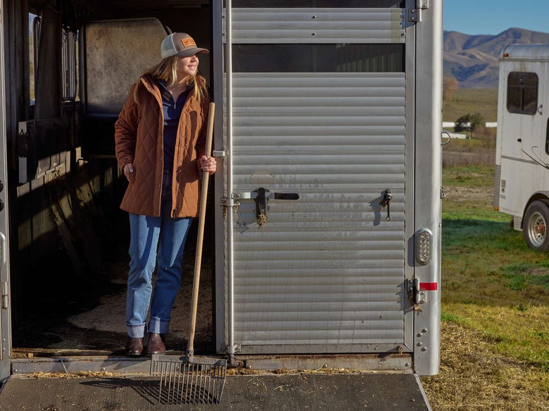 A woman holding a muck rake standing at the top of her horse trailer trailer ramp.