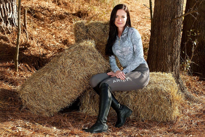 A woman sitting on a hay bale next to a tree, wearing the Ariat V Sport Tall Zip Boots.