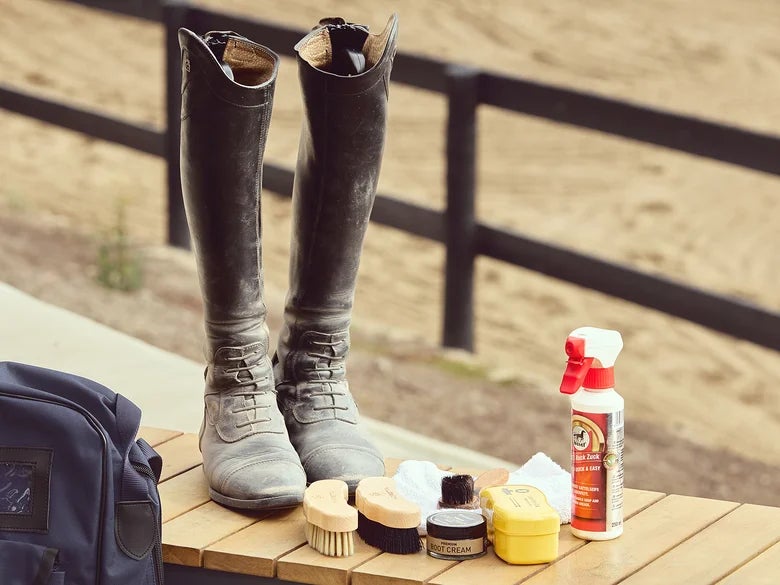 Dirty tall boots set on a wooden bench alongside all of the essential products to clean them and polish them. The wooden fence of an arena is in the background. 