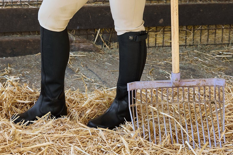 A rider standing in the hay with a manure fork in hand while wearing the Ovation Moorland II Highrider Women's Tall Boots in black. 