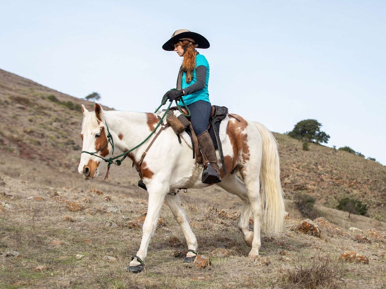 A trail rider riding her pinto horse across rocky terrain, with Explora Magic hoof boots on. 