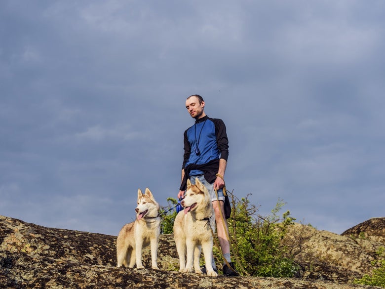 Male hiker standing on a rocky outcropping with two husky dogs on leashes. 