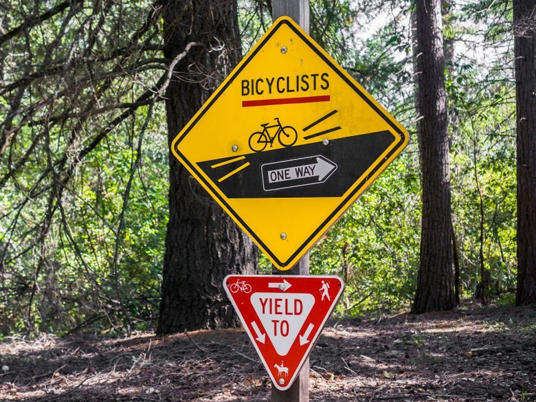 Two trail signs nailed to a wooden post. One larger, yellow sign is showing bicyclists need to go one way on the trails. The smaller, red sign is showing who has the right of way between hikers, bikers, and horseback riders.