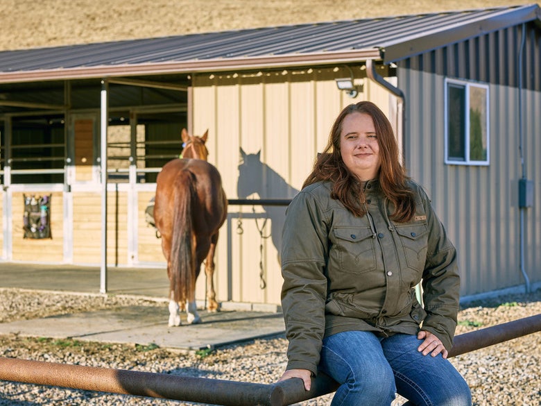 Woman sitting on fence in front of a barn wearing the Ariat Grizzly Parka 2.0 Jacket.