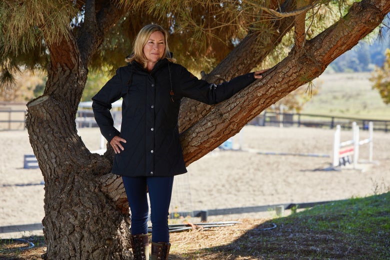 Woman standing in front of a tree outside of a riding arena wearing the Cinch Quilted Canvas Barn Coat Jacket.