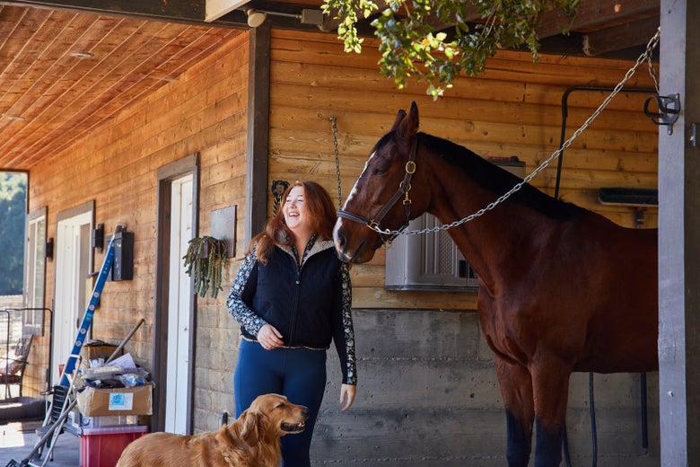 Woman standing with her bay horse and golden retriever at a wash rack wearing the Kerrits Double Up Reversible Vest.