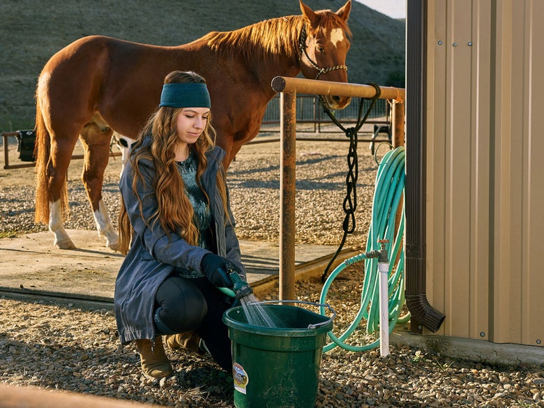 Woman filling up a bucket with water at a wash rack in front of a sorrel horse.