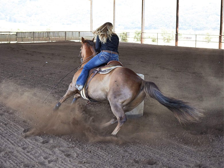 A lady wearing riding jeans and a black western top riding her sorrel horse around a barrel