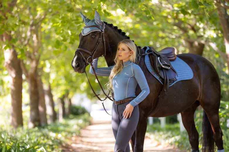 Woman standing with a dark bay horse in a driveway wearing a matching blue set from LeMieux.