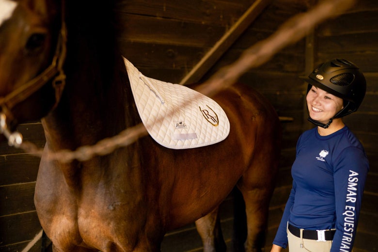 Smiling woman in riding apparel putting a saddle pad on her bay horse that's tied in crossties. 