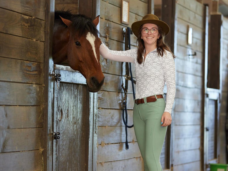 Rider petting a bay horse in a barn while wearing a Dacing Equestrian long sleeve, green Ariat breeches, and a brown belt and hat. 
