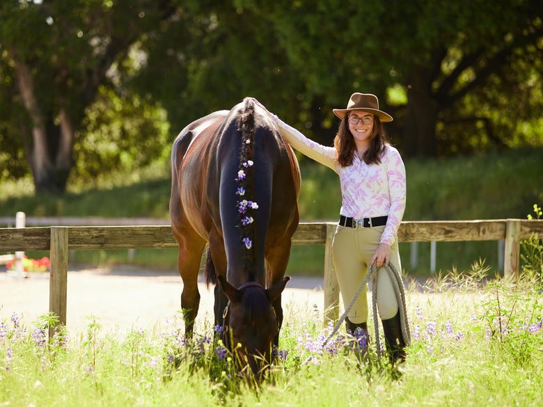 Women standing in a field of purple flowers, wearing her Kastel sun shirt, standing next to a bay horse with purple flowers in his mane.