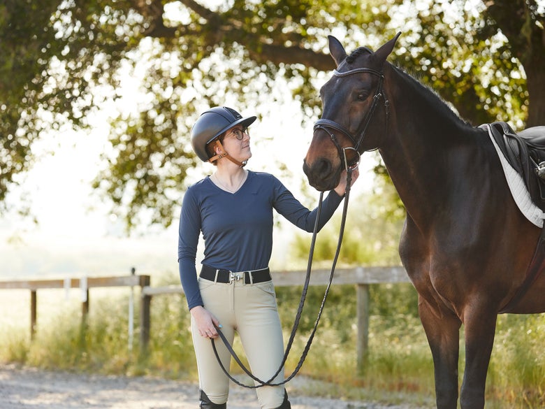 Rider wearing a long sleeve Tuffrider sun shirt in navy, holding the reins of her tacked up bay horse. 