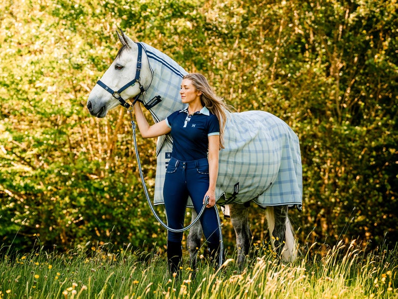 Girl with grey horse standing in a grassy field; horse is wearing the blue and white checkered Newmarket Horseware fly sheet.