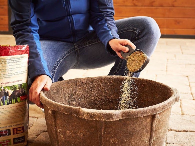 Person wearing a navy jacket and jeans pouring a powder joint supplement into a large black rubber grain tub.