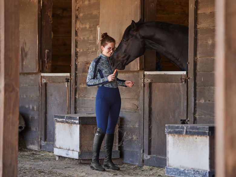 A rider feeding her bay horse a carrot while the horse is in the stall. 