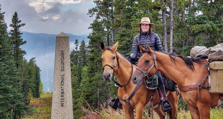 Gillian on horseback and ponying her pack horse, standing next to an international boundary statue. 