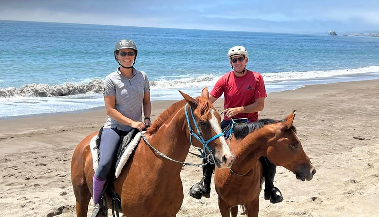 Heather and Jeremy Reynolds smiling at the camera on horseback, standing on the beach with a wave crashing in the background