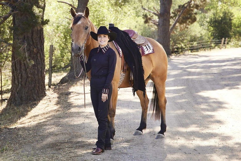 A Western rider dressed in black show attire with her dun horse. Her shirt cuffs and collar have a burgundy accent pattern that matches her horse's saddle pad. 
