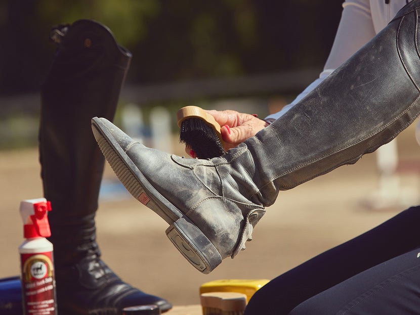 Woman demonstrating the first step of cleaning a tall boot, using a stiff brush to clean the dirt off of the boot. The second tall boot and other cleaning supplies are in the background. 