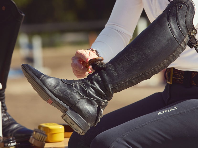 Woman showing the third step of cleaning a tall boot, using polish brush to apply boot cream polish to the boot. Her other cleaning supplies are next to her. 
