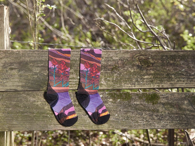The SmartWool Floral Field Wool Knee High Socks laying against a wooden fence. 