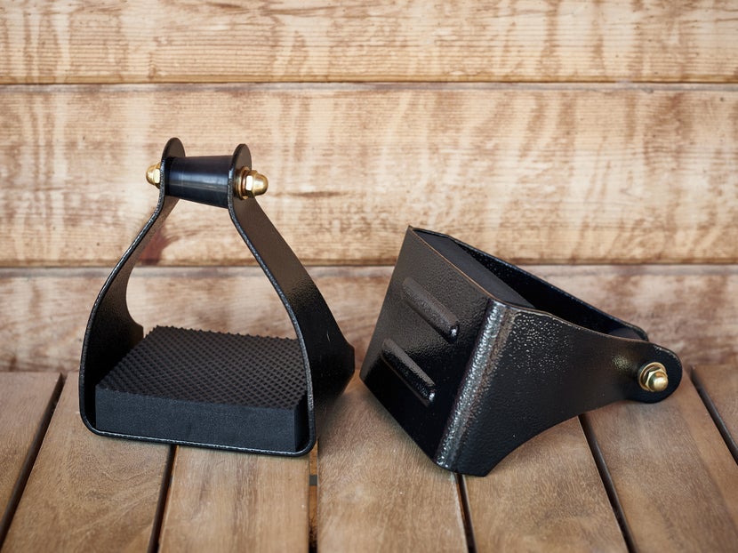 The Tucker Aluminum Trail Glide ErgoBalance Stirrups sitting on a wodden floor against a wooden barn. 