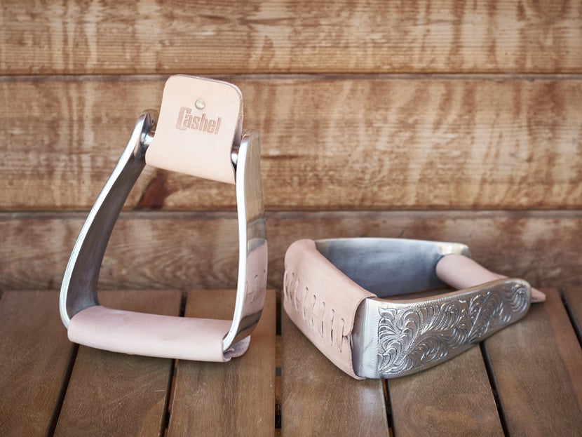 The Cashel Engraved Slanted Western Stirrups sitting on a wooden floor against a wooden building. 